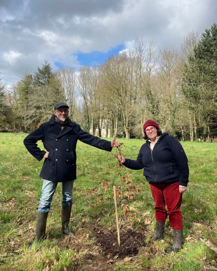 Deux personnes debout dans un champ au Manoir de Goaz Froment, tenant un jeune arbre planté. L'une porte un manteau noir et un chapeau, l'autre un bonnet rouge et un pull noir. Ciel nuageux en arrière-plan.
