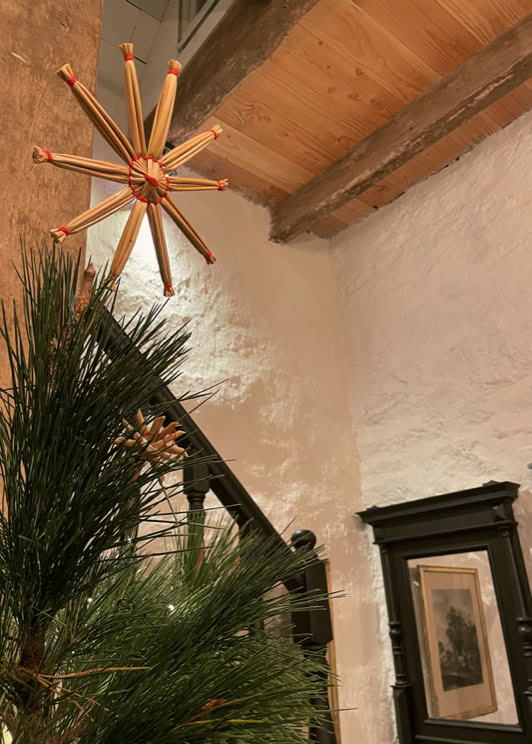 Décoration en forme d'étoile en bois suspendue près d'un sapin, avec un mur en pierre blanche et un plafond en bois en arrière-plan.