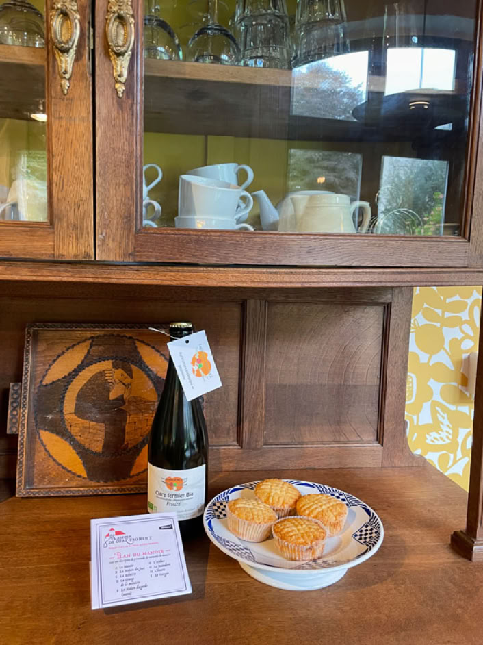 Bouteille de cidre bio, assiette de gâteaux bretons et document sur une buffet en bois devant un meuble avec vaisselle.