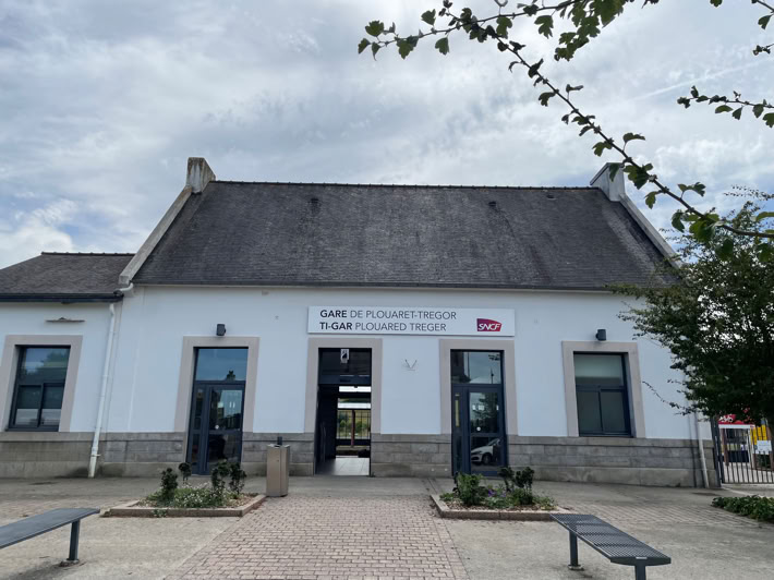 Bâtiment blanc (Gare TGV Plouaret-Trégor) avec toit en ardoise, trois portes, deux fenêtres, et un panneau au-dessus de l'entrée centrale. Arbres et bancs devant.