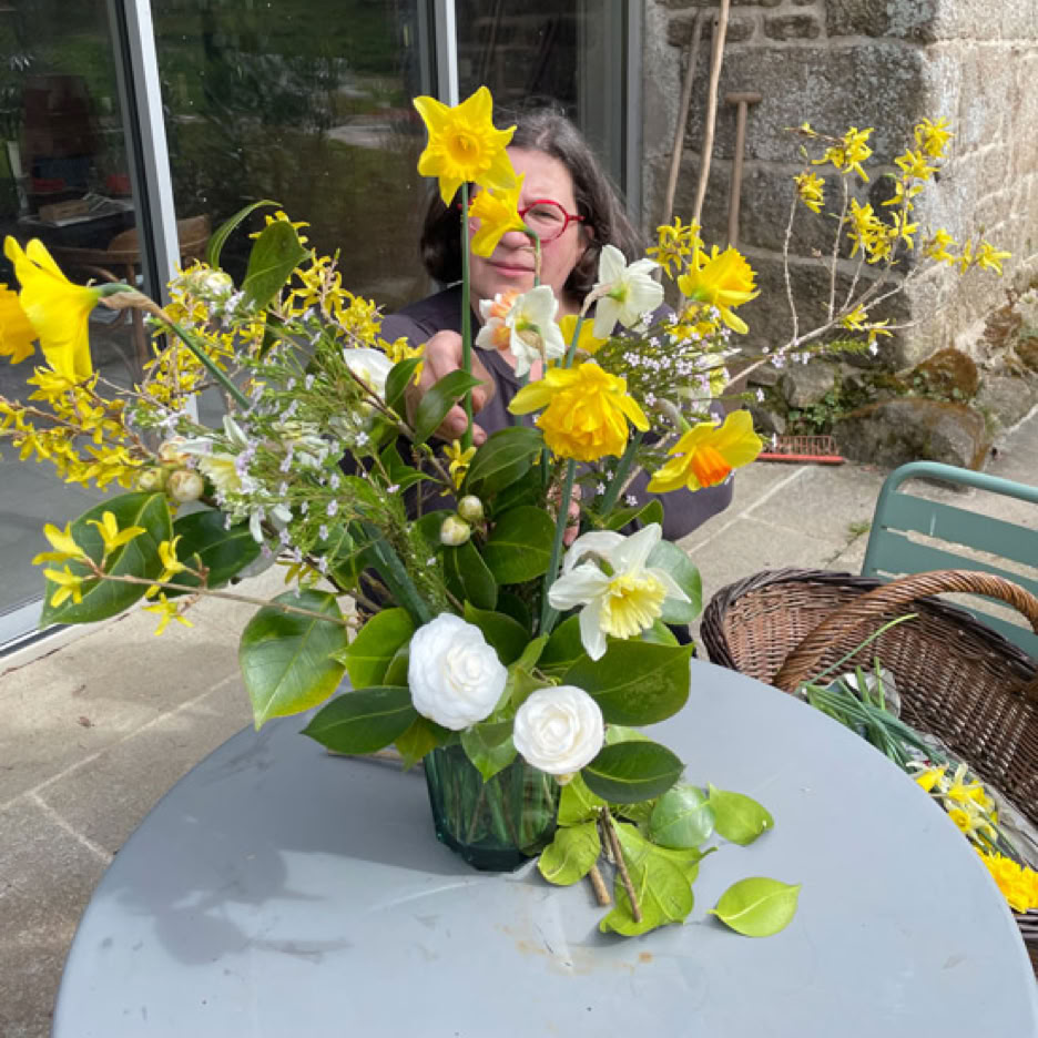 Un bouquet de fleurs jaunes et blanches sur une table grise, avec une personne en arrière-plan et une porte vitrée et un mur en pierre derrière.