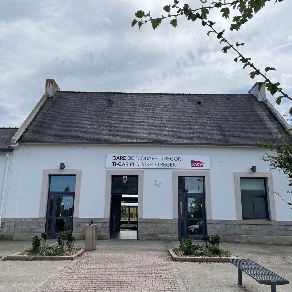 Bâtiment blanc (Gare TGV Plouaret-Trégor) avec toit en ardoise, trois portes, deux fenêtres, et un panneau au-dessus de l'entrée centrale. Arbres et bancs devant.