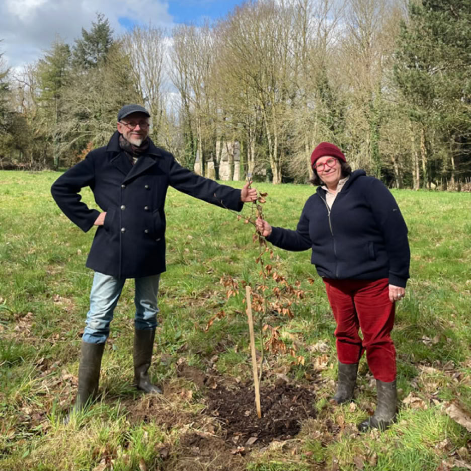 Deux personnes debout dans un champ au Manoir de Goaz Froment, tenant un jeune arbre planté. L'une porte un manteau noir et un chapeau, l'autre un bonnet rouge et un pull noir. Ciel nuageux en arrière-plan.