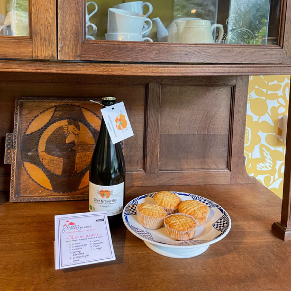 Bouteille de cidre bio, assiette de gâteaux bretons et document sur une buffet en bois devant un meuble avec vaisselle.