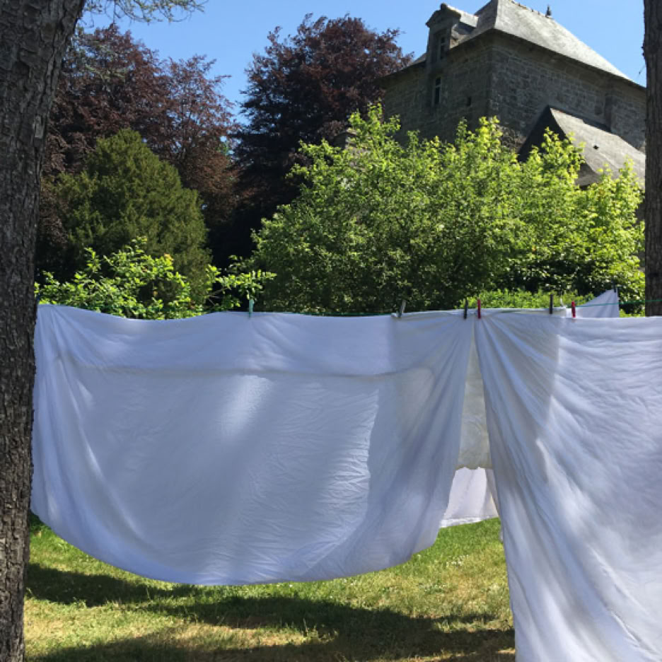 Draps blancs suspendus à une corde à linge entre deux arbres, avec un bâtiment en pierre et des arbres en arrière-plan.