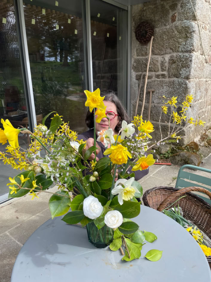 Un bouquet de fleurs jaunes et blanches sur une table grise, avec une personne en arrière-plan et une porte vitrée et un mur en pierre derrière.